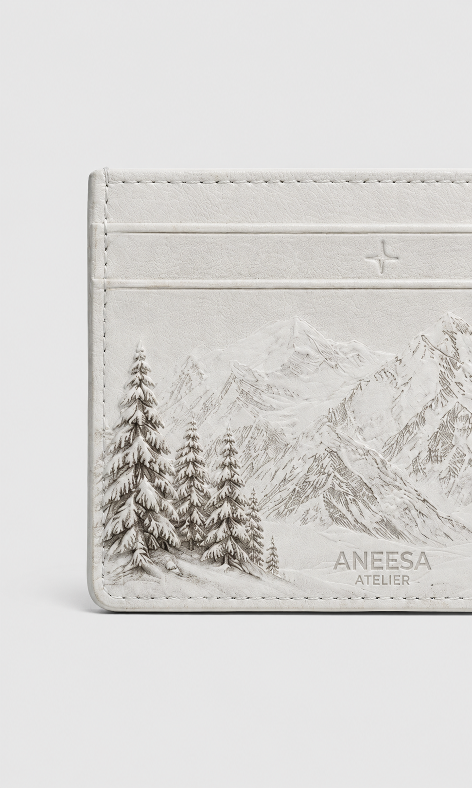 A close-up of a light beige leather card holder or wallet featuring an embossed scenic design of snowy pine trees and mountains, with the brand name 'ANEESA ATELIER' printed on the bottom right.