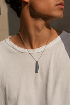 A close-up of a man wearing a silver-toned chain necklace with a matte gray-blue rectangular pendant, styled with a white crew-neck t-shirt.