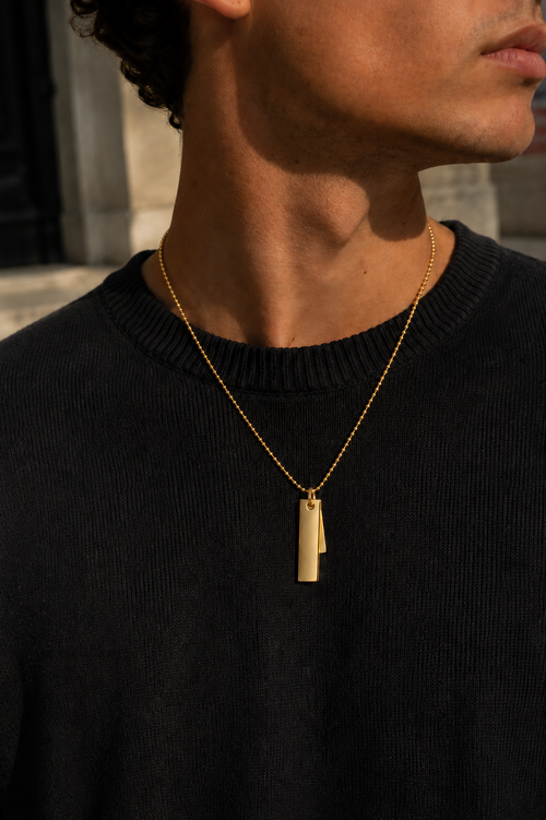 A close-up lifestyle shot of a man wearing a gold chain necklace with a polished rectangular bar pendant, styled with a black ribbed crewneck sweater.