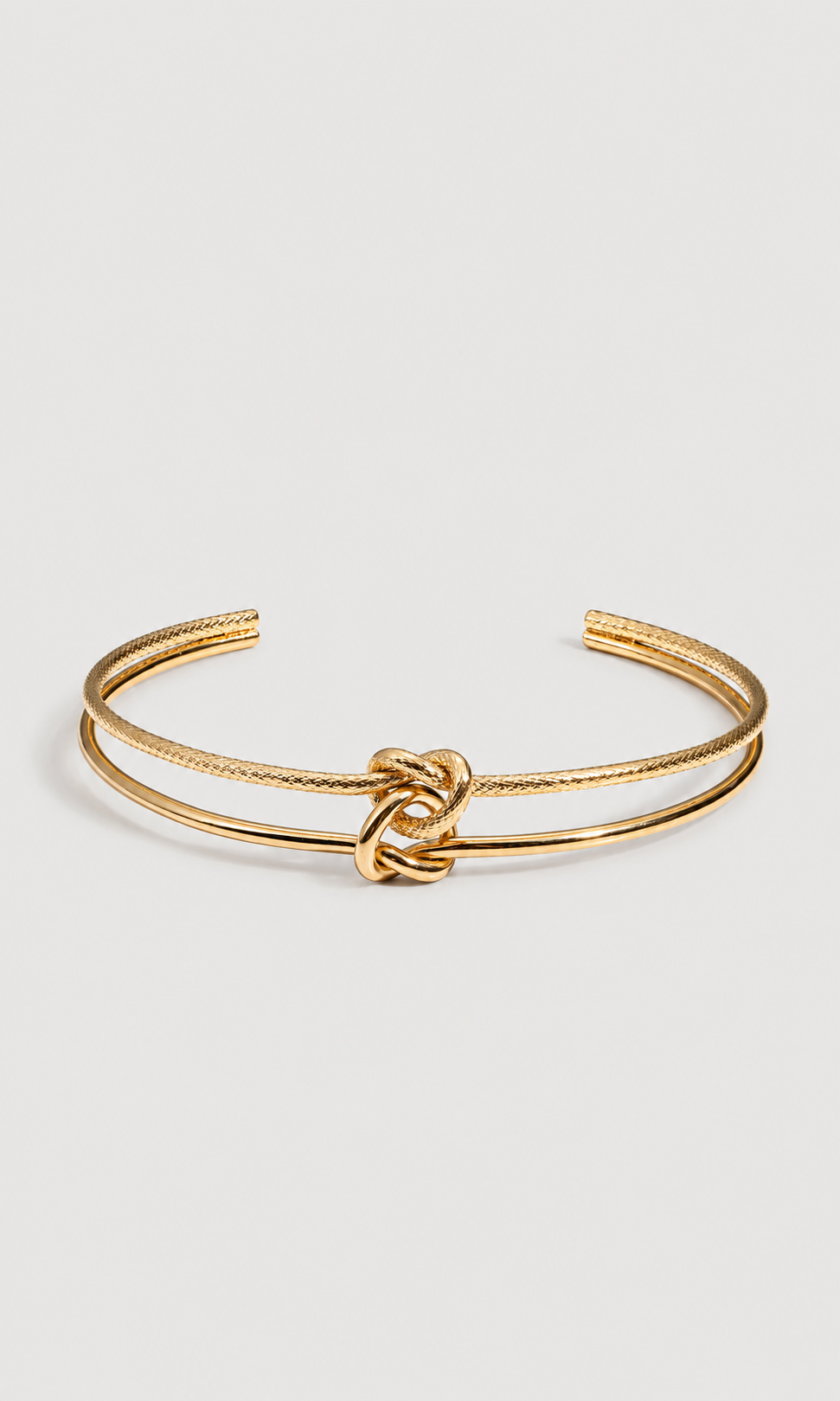 A gold-toned double-band cuff bracelet with a central knotted detail, shown on a light gray background.
