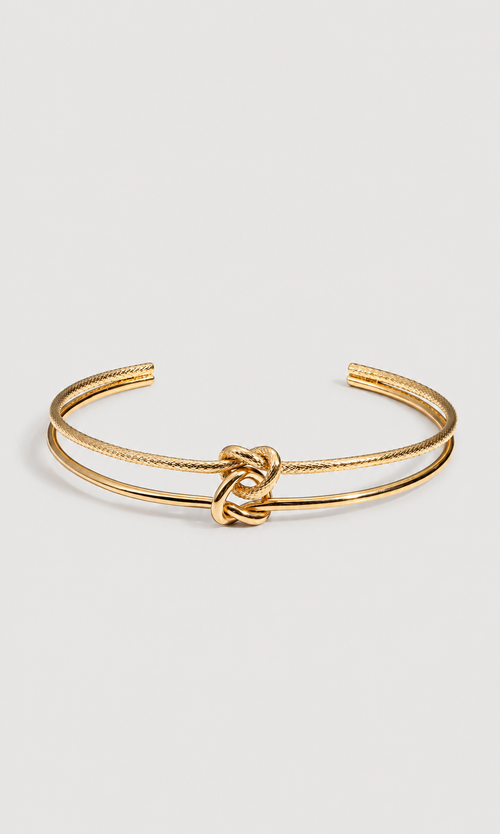 A gold-toned double-band cuff bracelet with a central knotted detail, shown on a light gray background.
