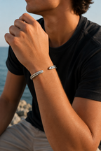 A close-up lifestyle shot of a man wearing a silver-toned braided bracelet on his wrist, set against a soft-focus background of the sea and sky.