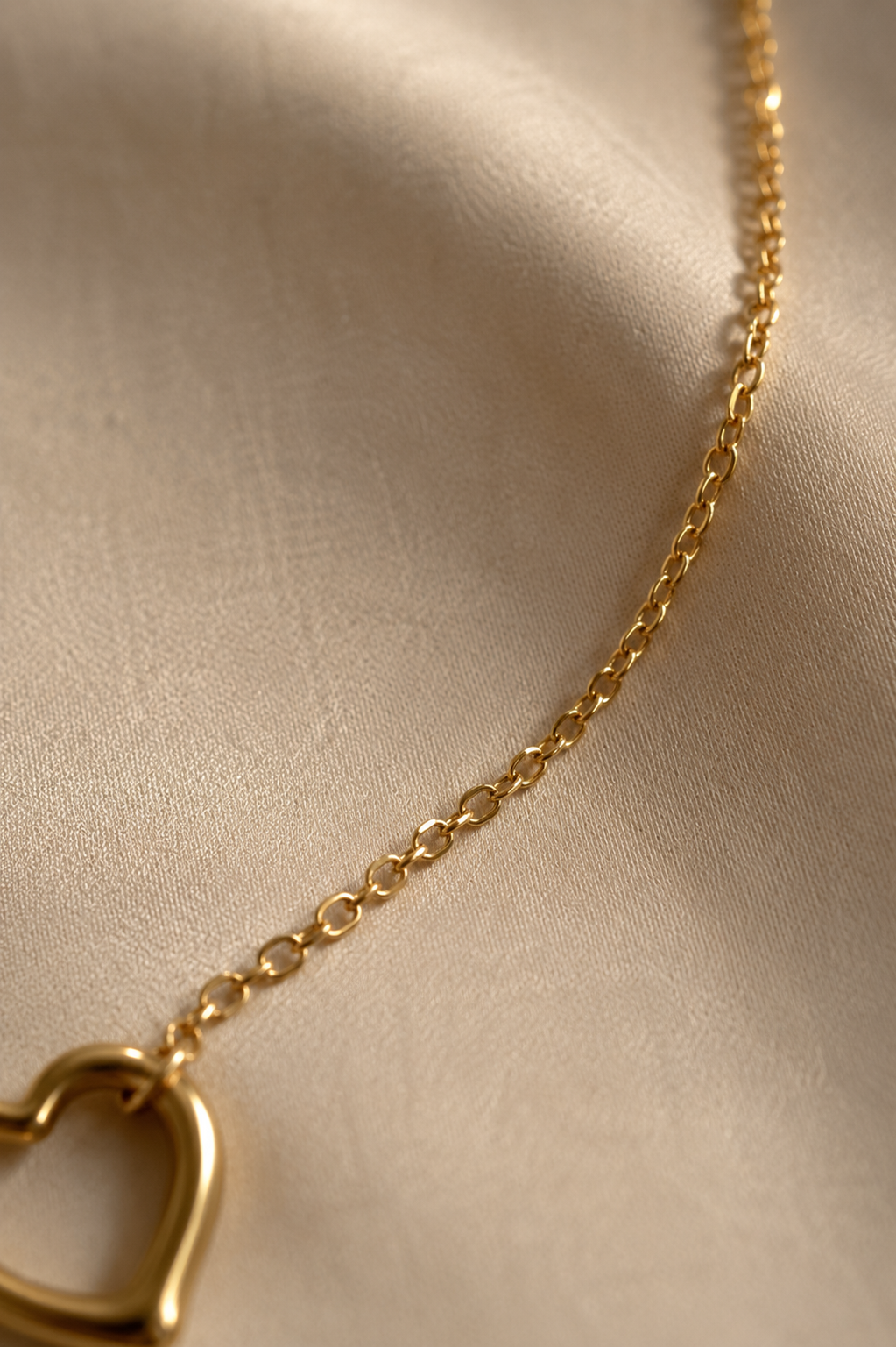 A close-up of a delicate gold chain necklace with a heart-shaped pendant, resting on a soft beige fabric background.