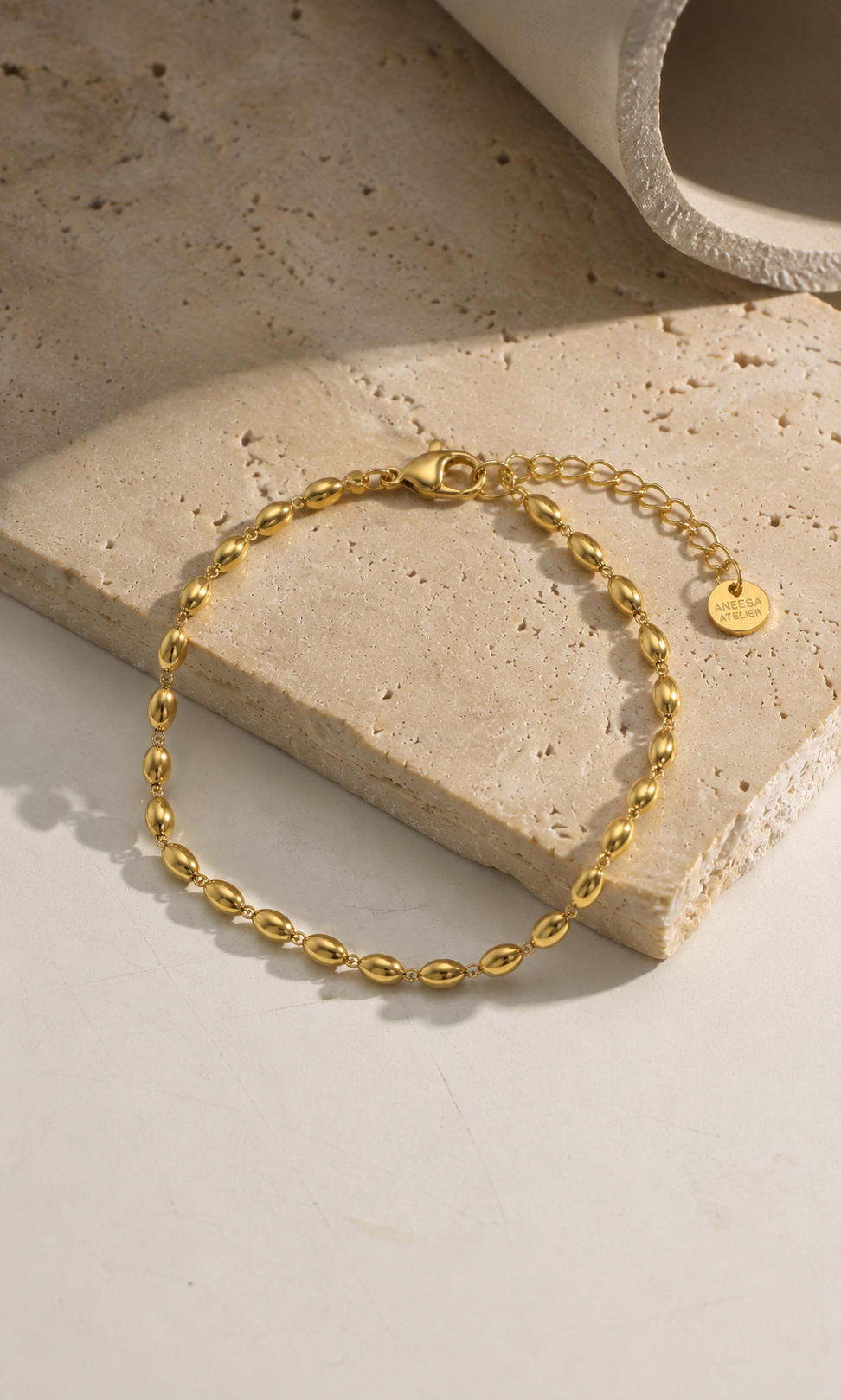 A gold chain bracelet with a small round tag, displayed on a textured beige stone slab against a light neutral background.