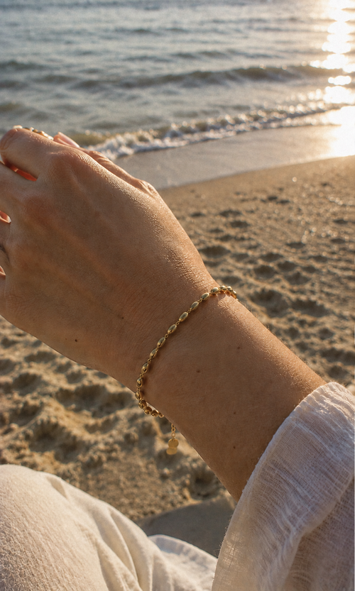 A close-up of a person's wrist wearing a delicate gold beaded bracelet, with a sandy beach and ocean waves in the background during golden hour.