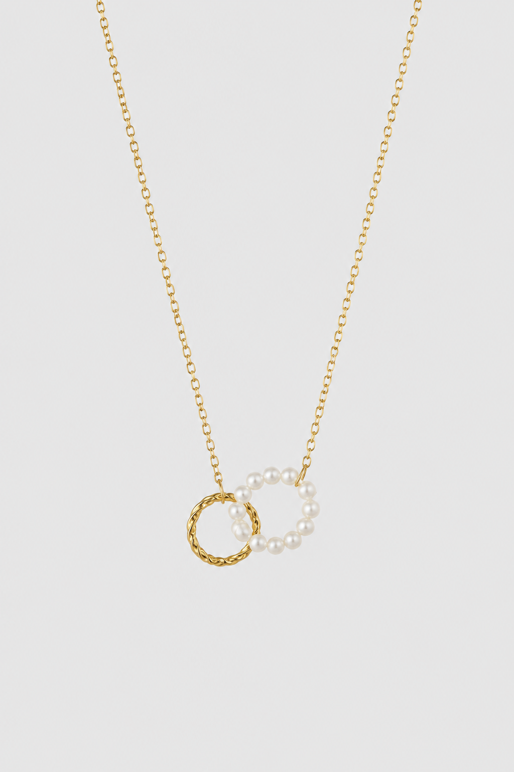 A delicate gold chain necklace featuring two interlocking circular pendants: one textured gold ring and one pearl-beaded gold ring, displayed against a solid light gray background.