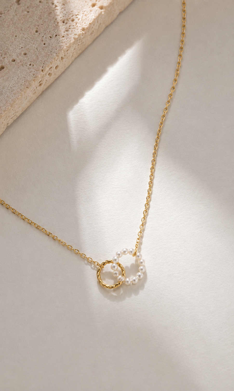 A delicate gold chain necklace with two interlocking ring pendants, one textured gold and one clear crystal, displayed on a warm beige surface with soft window light shadows.