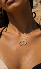 A close-up lifestyle shot of a model wearing a delicate gold necklace with two interlocking rings, one adorned with small white pearls and the other with tiny clear stones, set against a warm sandy background.