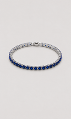 A tennis bracelet featuring rows of deep blue round-cut gemstones in a silver-toned setting, displayed on a neutral light gray background.