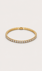 A gold tennis bracelet with a row of clear gemstones along the front, displayed on a light beige background.