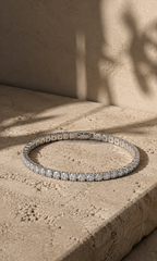Silver tennis bracelet with sparkling stones, photographed on a warm beige natural stone surface in soft sunlight.