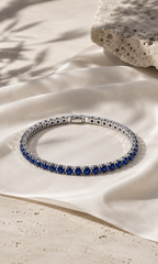 Silver tennis bracelet with sparkling stones, photographed on a warm beige natural stone surface in soft sunlight.