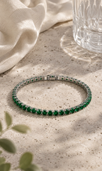Green gemstone tennis bracelet in silver setting, styled on a textured beige stone surface with soft sunlight, linen fabric and subtle greenery.
