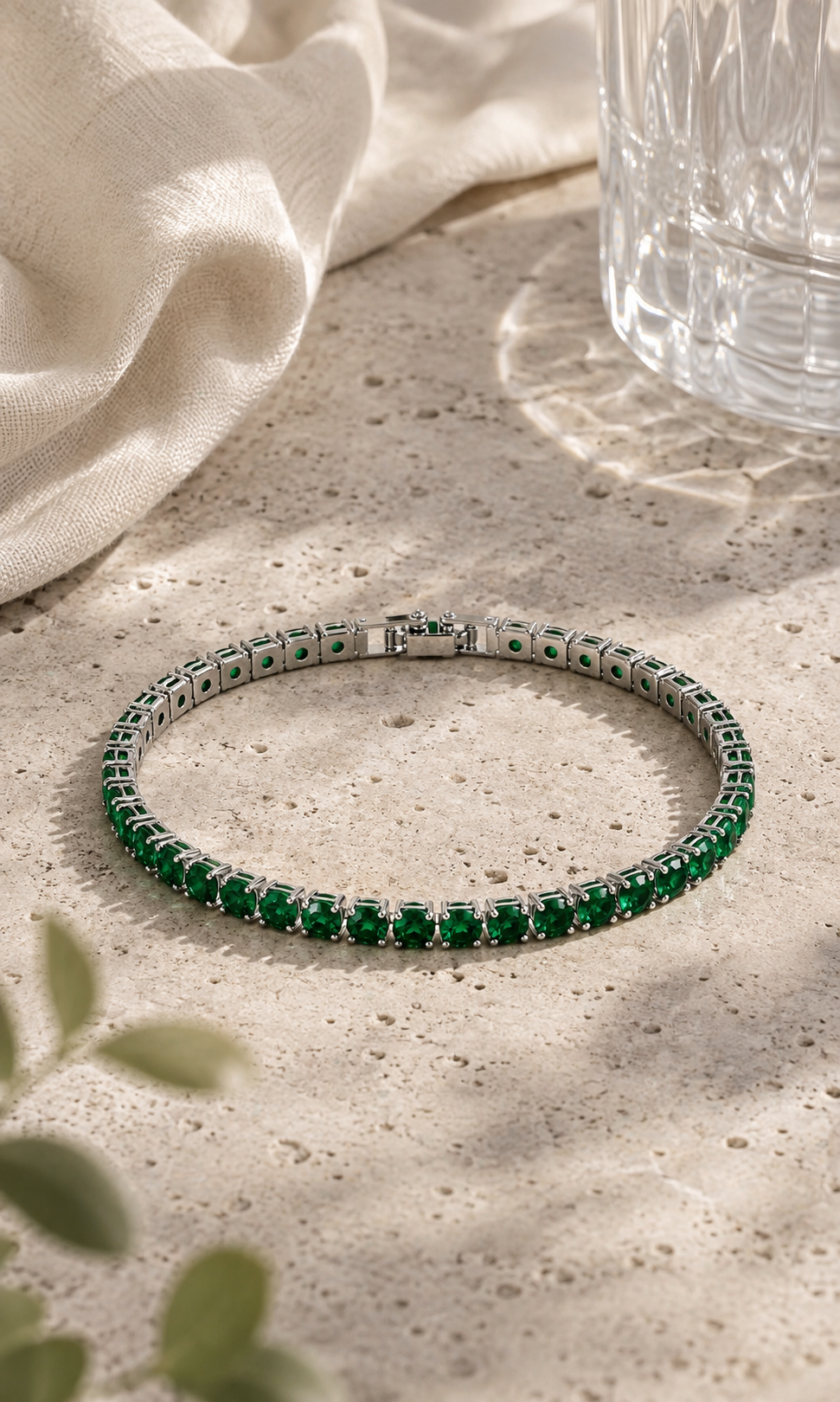 Green gemstone tennis bracelet in silver setting, styled on a textured beige stone surface with soft sunlight, linen fabric and subtle greenery.