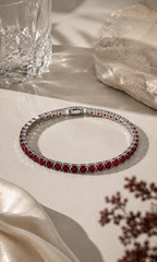 Ruby-red gemstone tennis bracelet with a silver setting, styled on a warm beige surface with soft sunlight, ivory fabric, natural stone and elegant decorative details.