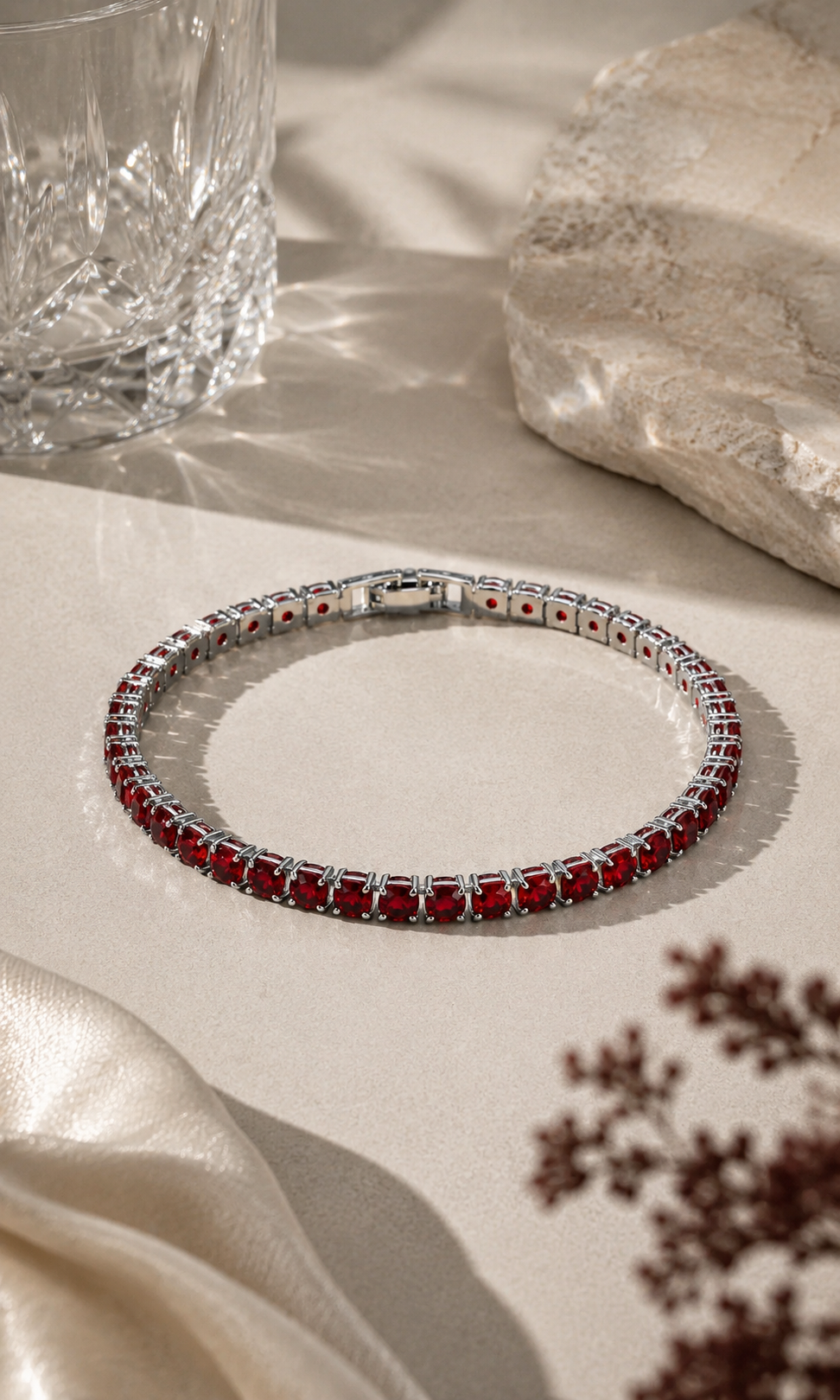 Ruby-red gemstone tennis bracelet with a silver setting, styled on a warm beige surface with soft sunlight, ivory fabric, natural stone and elegant decorative details.