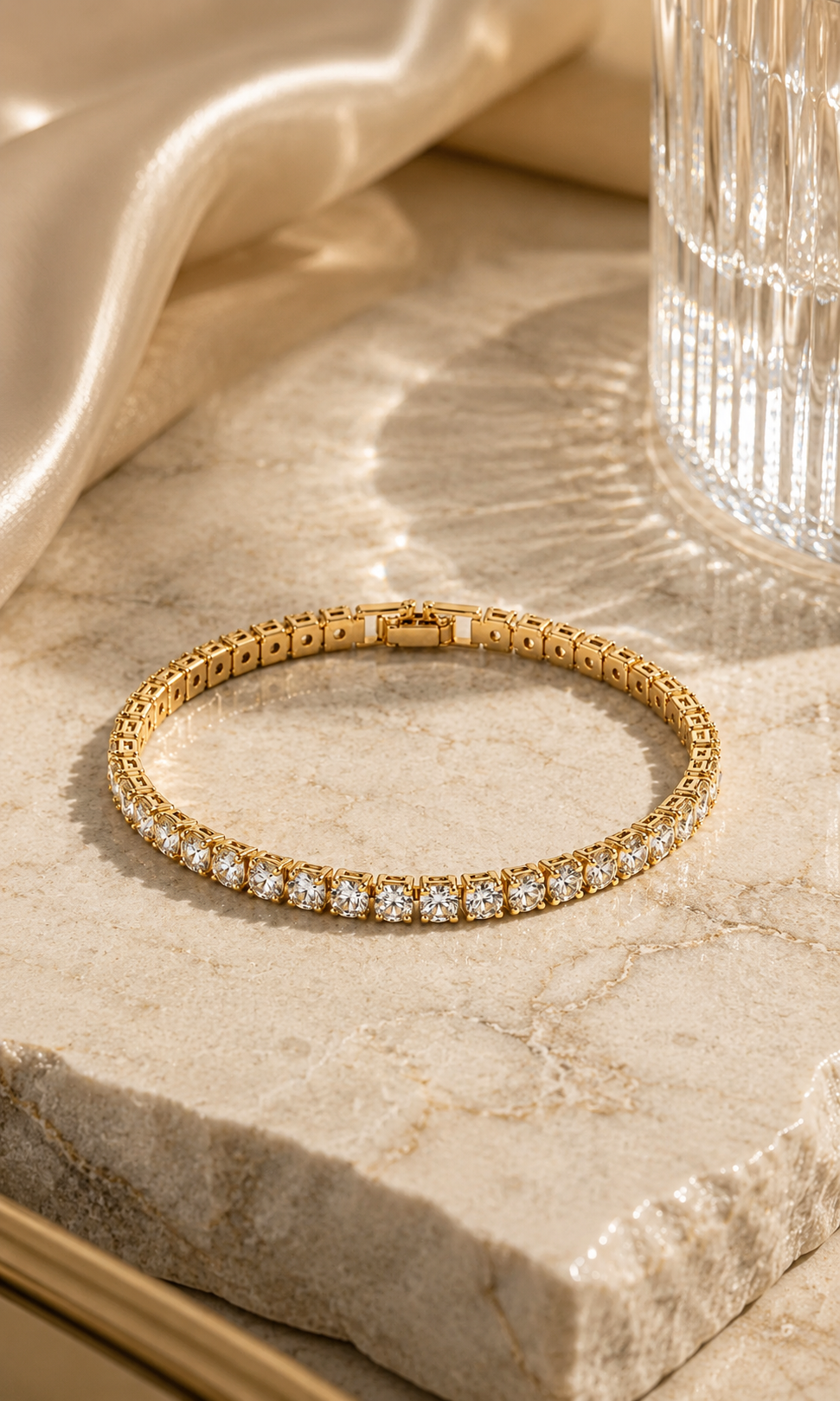Gold tennis bracelet with sparkling clear gemstones, styled on beige marble with satin fabric and warm luxury lighting.