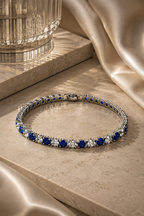 Blue and clear gemstone tennis bracelet in silver setting, styled on beige marble with champagne satin and warm luxury lighting.