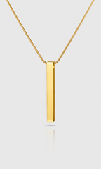 A gold bar pendant necklace on a delicate gold chain, shown against a light gray background with a soft reflection below.