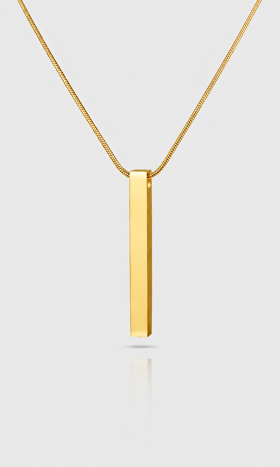 A gold bar pendant necklace on a delicate gold chain, shown against a light gray background with a soft reflection below.