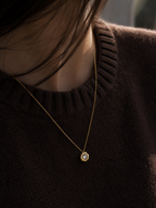 A close-up lifestyle shot of a person wearing a delicate gold chain necklace with a small round pendant featuring a clear central stone and gold accents, set against a dark brown knit sweater.