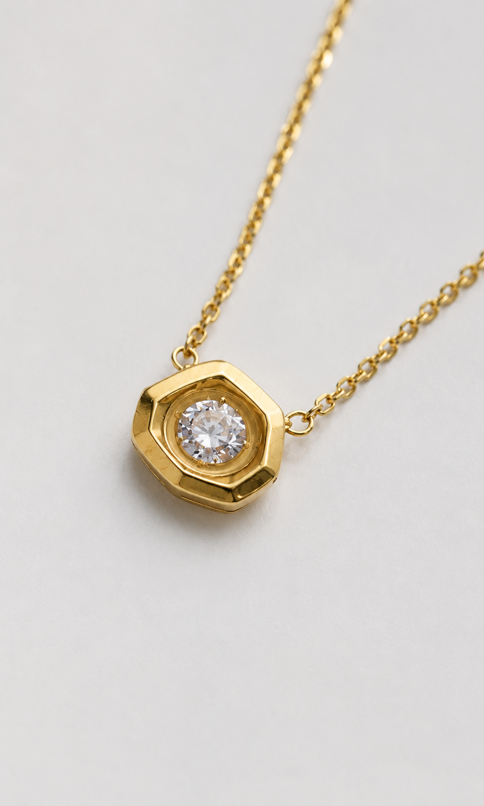 A gold necklace with a faceted octagonal pendant featuring a clear round gemstone, displayed on a light gray background.