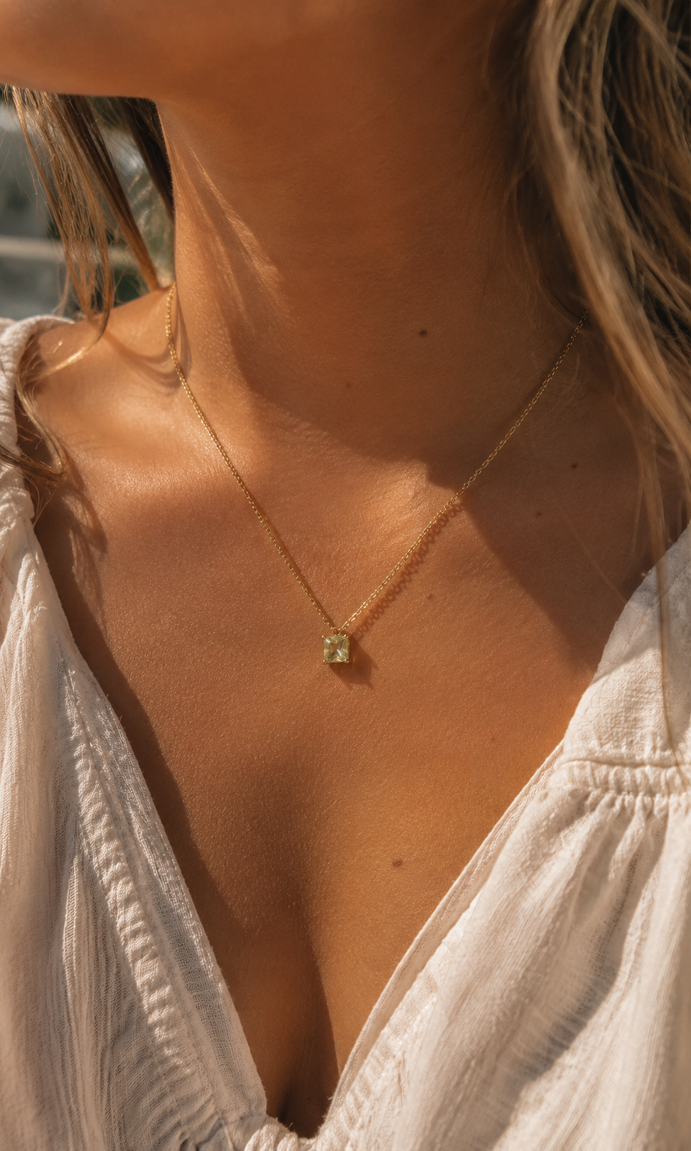 A close-up lifestyle shot of a woman wearing a delicate gold chain necklace with a small square-cut green gemstone pendant, styled with a white top under warm sunlight.