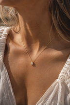 Elegant gold necklace with a square-cut clear gemstone pendant worn on a woman’s neck and collarbone, styled with a textured white blouse in warm natural sunlight.