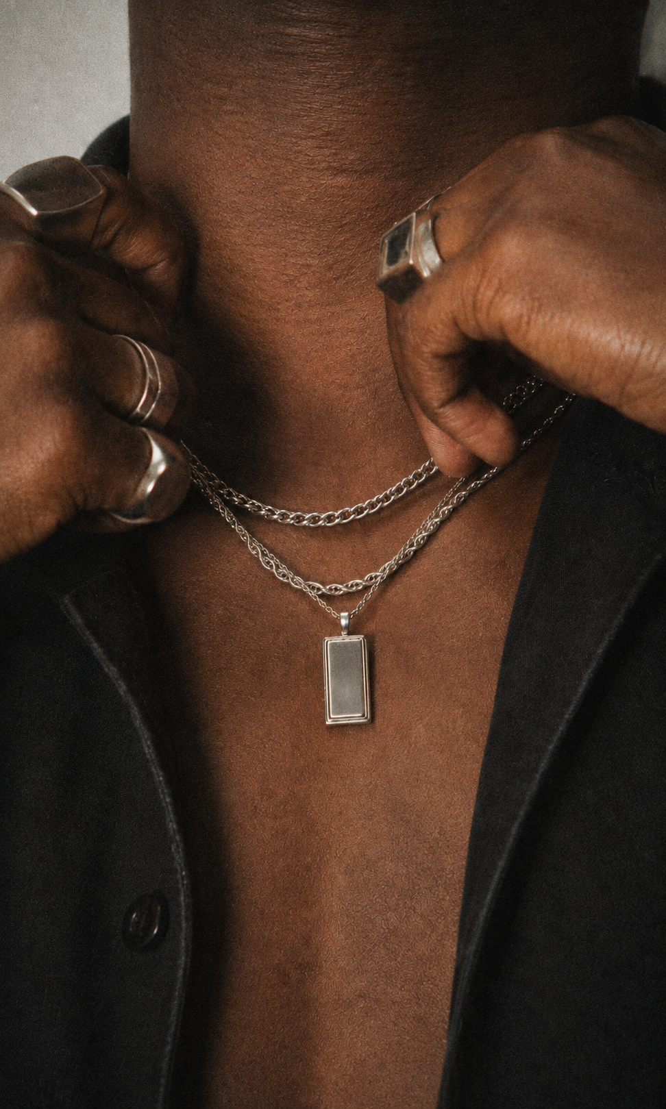 A close-up of a person wearing a layered silver necklace with a rectangular pendant, styled with silver rings, over a black shirt.