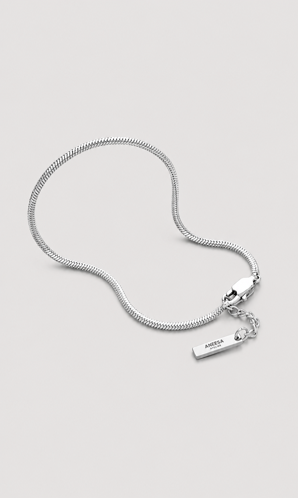 A silver snake chain necklace with a small rectangular metal tag engraved with the brand name 'ANEESA ATELIER', displayed on a light gray background.