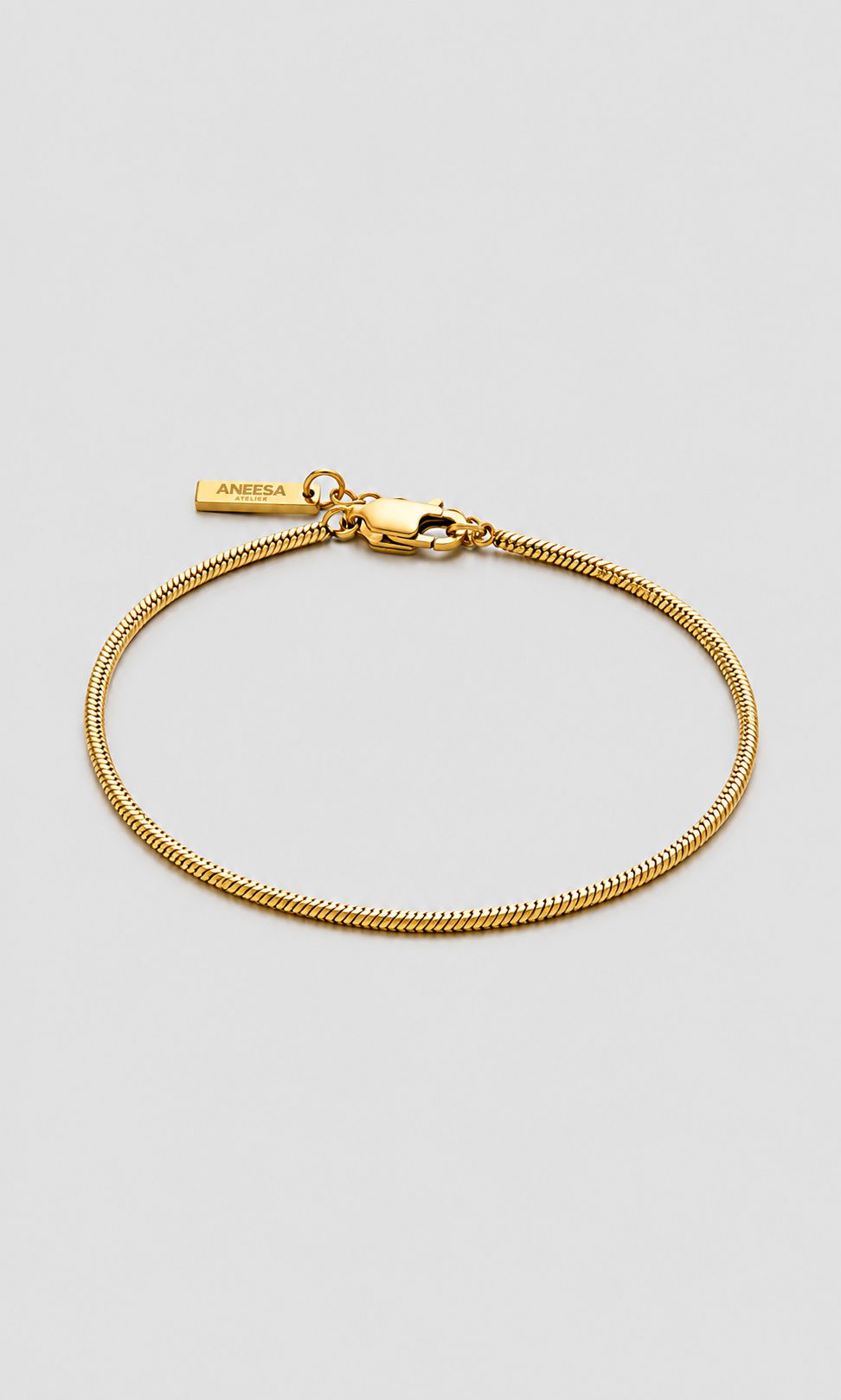 A gold chain bracelet with a lobster clasp and a small branded tag reading 'ANEESA ATELIER' on a light gray background.