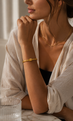 A woman wearing a delicate gold bangle bracelet with a small central charm, styled with a gold chain necklace and gold hoop earring, wearing a white linen shirt.