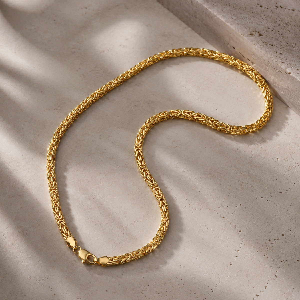 A gold chain necklace with a braided, rope-like link pattern, displayed on a textured light beige stone surface with soft shadows.